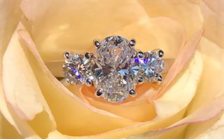 A diamond ring with a central oval diamond flanked by two smaller round diamonds on each side, set in a silver band, is nestled inside the petals of a yellow rose.