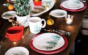 A festive table setting with a small decorated Christmas tree centerpiece. The table features plates with green leaf patterns, a red bowl, a mug, candles, cutlery wrapped in gold ribbons, and scattered oranges, creating a holiday atmosphere.