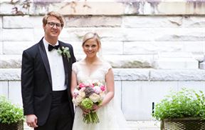 A bride and groom stand side by side, smiling and dressed in wedding attire. The bride is holding a colorful bouquet of flowers and wearing a white gown. The groom is wearing a black tuxedo and glasses. They pose in front of a light-colored stone wall with greenery on both sides.