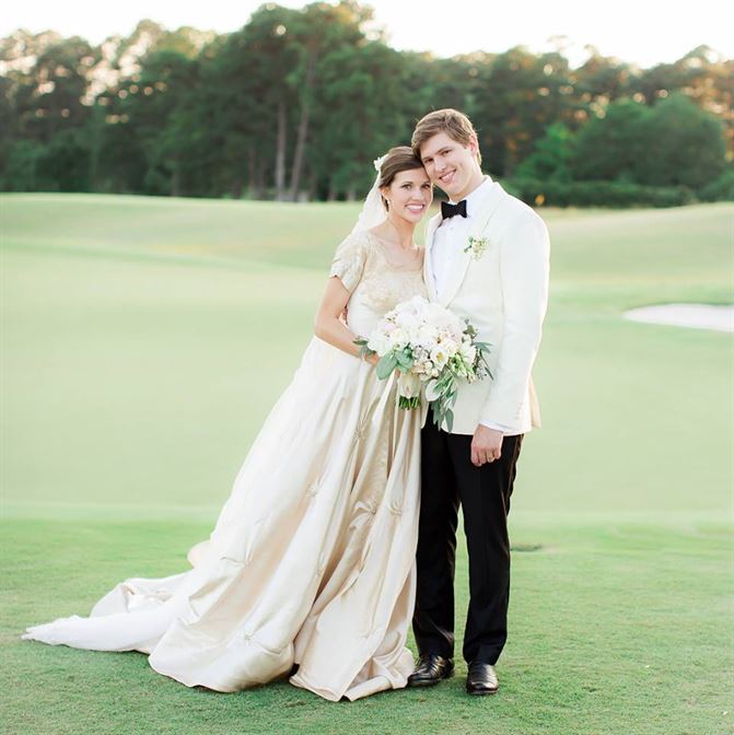 A bride and groom stand closely together on a grassy field, smiling for the camera. The bride wears an elegant white dress and holds a bouquet of white flowers, while the groom is dressed in a white tuxedo with black pants. Trees are visible in the background.