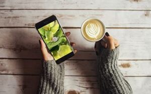 A person wearing a gray sweater holds a smartphone in their left hand displaying an image of green flowers and a diamond ring. In their right hand, they hold a cup of latte with a heart-shaped foam art, all placed on a rustic wooden table.