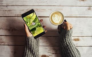 A person wearing a gray sweater holds a smartphone in their left hand displaying an image of green flowers and a diamond ring. In their right hand, they hold a cup of latte with a heart-shaped foam art, all placed on a rustic wooden table.