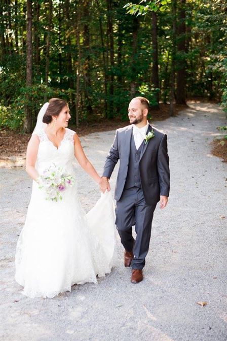 A bride and groom walk hand in hand along a wooded path. The bride wears a white gown, holding a bouquet of flowers, and the groom wears a grey suit with a white shirt. Both are smiling and looking at each other lovingly. The background is filled with trees and greenery.