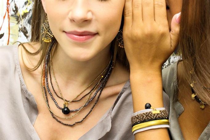 Two women are close together; one whispers to the other. The woman in front has light skin, wears multiple intricate necklaces and dangly earrings, and sports a neutral-toned blouse. The woman's arm in the background shows bracelets with various designs.