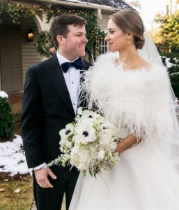 A bride and groom stand together in an outdoor winter setting. The bride is in a white wedding gown with a feathered wrap, holding a bouquet of white flowers. The groom wears a black tuxedo with a blue bow tie. They are facing each other and smiling.