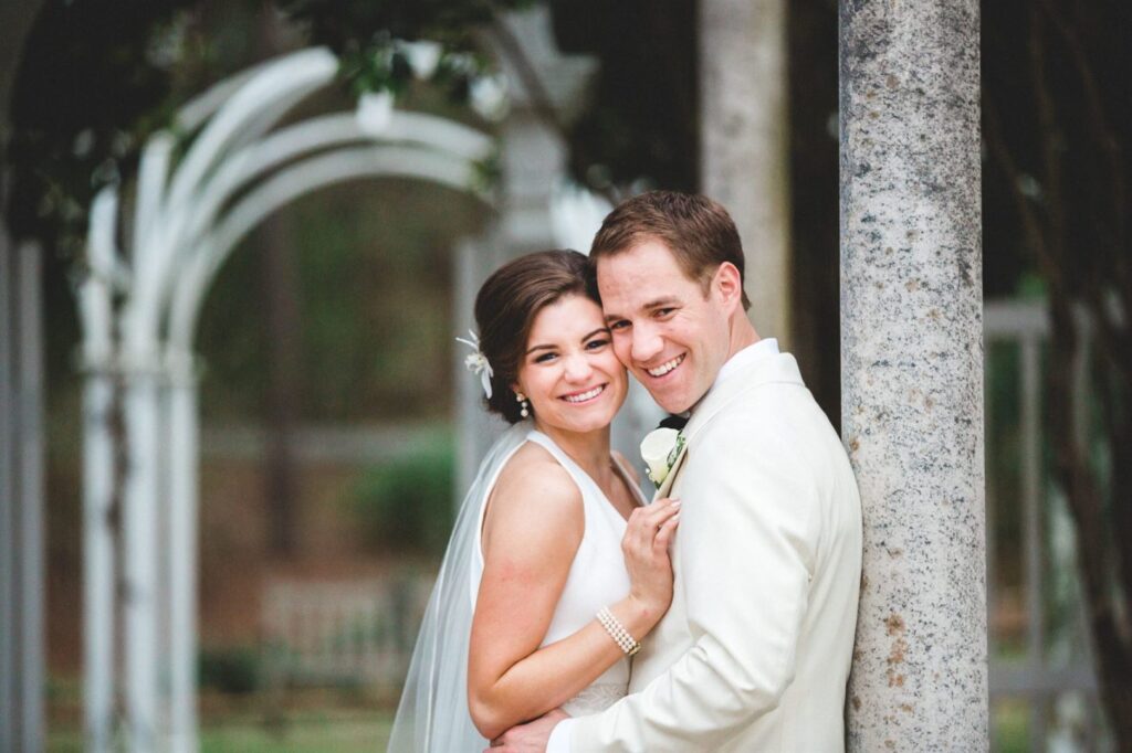 A bride and groom are embracing and smiling under an outdoor archway. The bride is wearing a white dress and veil, while the groom is dressed in a white suit. They stand next to a stone pillar, with greenery and another archway visible in the background.