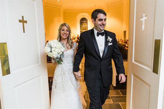 A bride and groom hold hands and smile as they walk through open double doors adorned with cross symbols, into a well-lit room. The bride wears a white, floral lace dress and carries a bouquet of white flowers, while the groom is dressed in a black tuxedo.