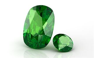 Two green emerald gemstones, one larger and oval-shaped and the other smaller and round, placed on a white background. Both gems have a brilliant cut, displaying facets that reflect light.