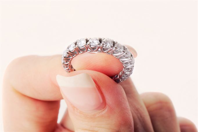 A hand with neatly manicured nails holds a silver ring adorned with several small, sparkling diamonds set in a lace-like pattern. The ring is being displayed against a plain, light-colored background.