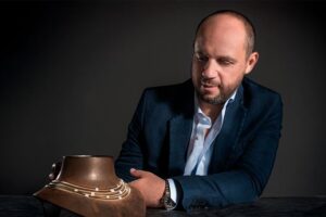 A man with a beard, wearing a suit, gazes intently at a bronze, ancestral-like artifact on a table. The artifact has several concentric layers or rings around it. The background is dark, emphasizing the man and the object.