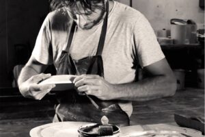 A person wearing a gray shirt and a dark apron is carefully inspecting a plate in a workshop. The scene includes various pottery tools and materials on the table, suggesting a focus on ceramic craftsmanship.