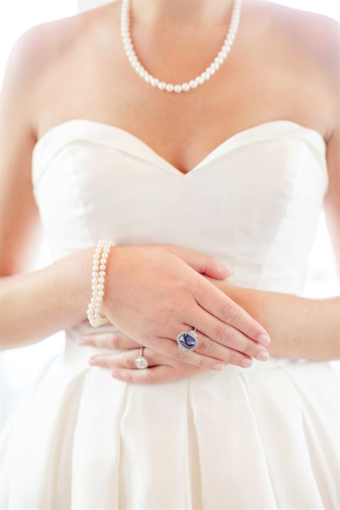 A bride in a strapless white wedding dress stands with her hands delicately clasped. She wears a pearl necklace, a pearl bracelet, a gemstone ring on her middle finger, and a wedding ring on her ring finger. The background is softly blurred.