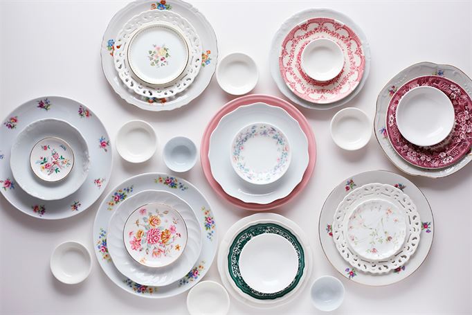 Assorted porcelain plates and bowls with various floral and decorative patterns are arranged on a white surface. The tableware features different sizes, designs, and colors including pink, red, green, and blue accents.