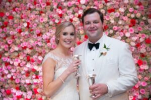 A bride and groom dressed in a white wedding dress and white tuxedo stand smiling, each holding a silver goblet. They are in front of a wall covered with numerous pink and white roses.