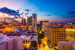 Panoramic view of a city skyline at dusk, featuring illuminated high-rise buildings and streets lined with trees. The sky transitions from deep blue to light orange as the sun sets, creating a vibrant, colorful backdrop with scattered clouds.