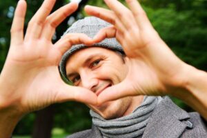 A smiling man wearing a grey knit hat and scarf forms a heart shape with his hands in front of his face. The background is green and leafy, suggesting he is outdoors.