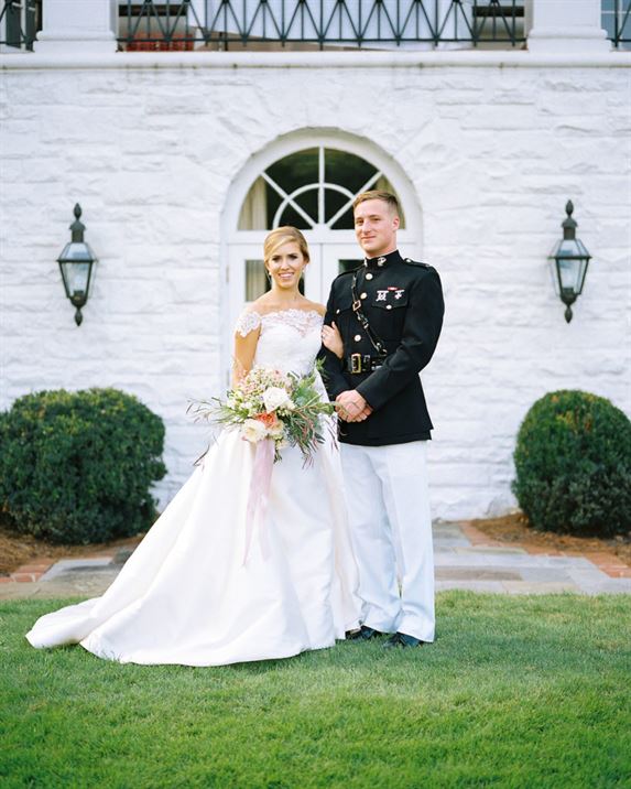 A bride and groom pose for a wedding photo in front of a white stone building. The bride wears an off-the-shoulder white gown and holds a floral bouquet. The groom, in a formal military uniform, stands beside her. Two wall lanterns flank the doorway behind them.