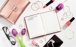 An open notebook with "Goals for the week" written on the top is surrounded by a rose gold purse, glasses, pink lipstick, pink nail polish, gold jewelry, a smartphone with earphones, a pen, and purple tulips scattered on a white surface.