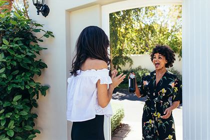 Two women are greeting each other at an open gate in a garden. One woman, wearing a white off-shoulder top, is facing the other woman, who is holding a wine bottle and wearing a black floral dress with a surprised and joyful expression.