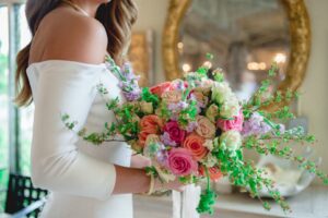 A bride in an off-the-shoulder white wedding dress holds a vibrant bouquet of pink, purple, and white flowers accented with greenery. A large antique mirror and soft interior decor are blurred in the background.