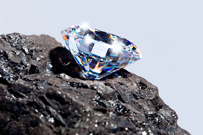 A sparkling diamond rests on a piece of dark, rough rock. The diamond's facets reflect light, creating a colorful spectrum that contrasts with the dull, textured surface of the rock. The background is a soft, light blue gradient.