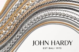 Various intricately designed gold and silver chain bracelets cascading across a white background with the text "John Hardy, Est Bali 1975" at the bottom.