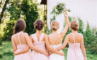 Four women stand with their backs to the camera, arms linked around each other. Three wear light pink dresses and the fourth, in white, holds a bouquet of flowers up in the air. They are outdoors with greenery in the background.