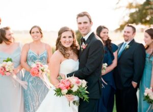 A bride and groom stand smiling and embracing in the center of a group of bridesmaids and groomsmen. The bride wears a white strapless dress and holds a colorful bouquet, while the groom is in a black suit and bowtie. The background is an outdoor setting.