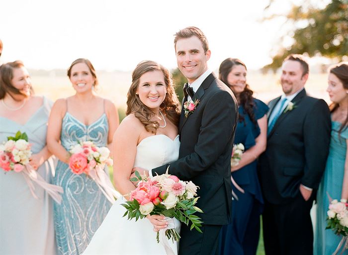 A bride and groom stand smiling and embracing in the center of a group of bridesmaids and groomsmen. The bride wears a white strapless dress and holds a colorful bouquet, while the groom is in a black suit and bowtie. The background is an outdoor setting.