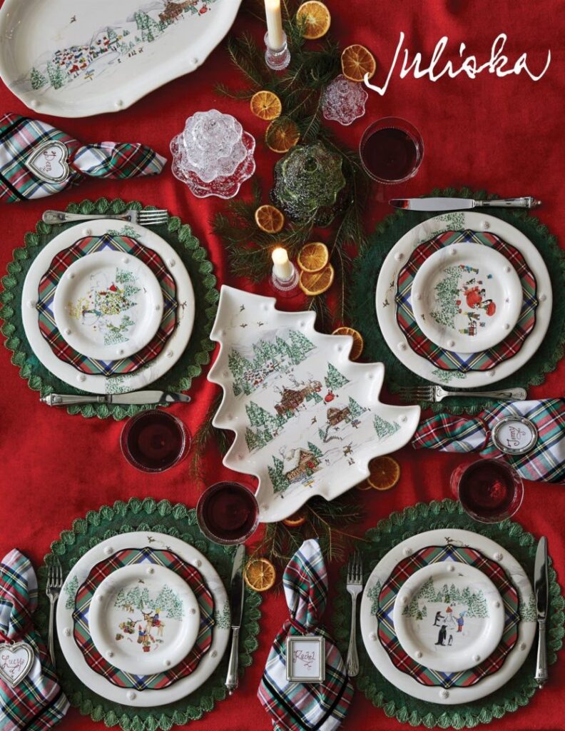 Festive dining table set with Juliska holiday-themed dishware featuring Christmas scenes. The plates are arranged on red and green plaid mats, with green doilies underneath. Pine branches and dried orange slices accent the centerpiece, with lit candles.