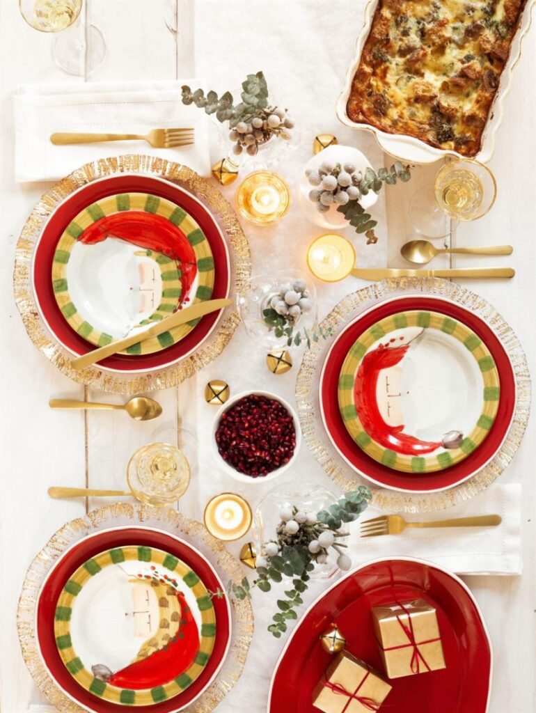 Festive dining table set with colorful plates, gold cutlery, candles, and sprigs of eucalyptus. A casserole dish is placed at the top, with a bowl of pomegranate seeds and three small wrapped gifts beside the table settings.