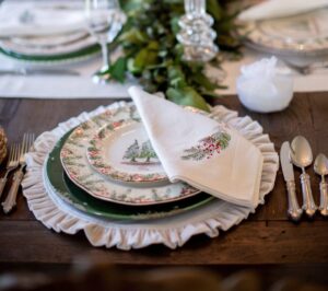 A festive table setting with layered plates featuring holiday designs, including a Christmas tree and holly. A folded napkin with embroidered pine cones and greenery is placed on top. Silverware and a wine glass are neatly arranged nearby. A leafy garland decorates the center.