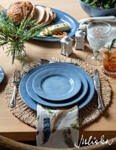 A place setting with blue plates on a woven placemat, a printed napkin tucked under the plate, silver cutlery, a crystal glass, and an orange cocktail. Bread, cheese, and a salad are in the background. The logo "Juliska" is in the bottom right corner in white text.