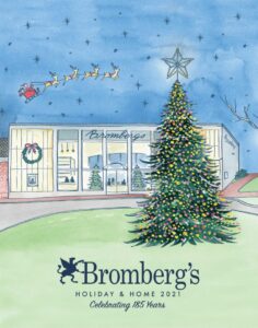 Illustration of a store with "Bromberg's" signage, decorated for the holidays. A large Christmas tree with colorful lights stands prominently in front. Santa and his reindeer fly across a starry sky above. Text reads "Bromberg's Holiday & Home 2021 Celebrating 185 Years.