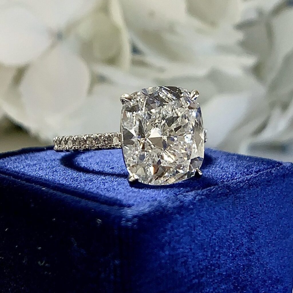 A close-up image of a sparkling diamond engagement ring. The ring features a large, cushion-cut diamond set on a silver band adorned with smaller diamonds. The ring is placed on a blue velvet surface with a blurred white floral background.