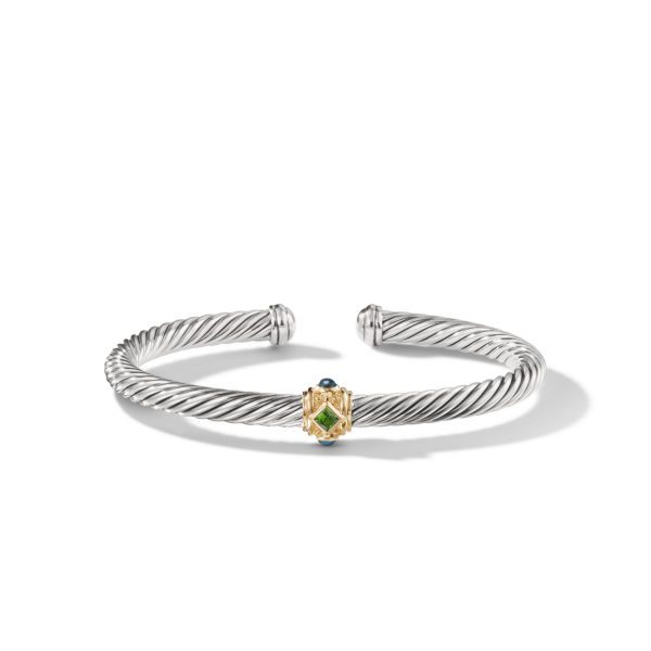 A silver twisted cuff bracelet with open ends features a small gold-accented centerpiece adorned with two gemstones – one green and one blue. The design is elegant and minimalist, highlighting the twisted rope texture and the contrasting colors of the stones.