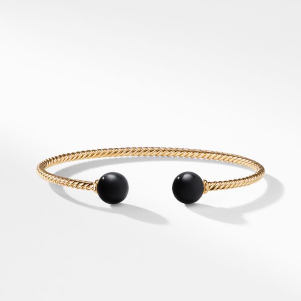 A gold twisted cable cuff bracelet with black spherical ends is shown against a plain white background. The bracelet is slightly open, highlighting its elegant and minimalist design.