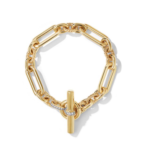 A gold chain bracelet with elongated rectangular links and a cylindrical toggle clasp that features small embedded gemstones. The bracelet is arranged in a circular shape against a white background, with shadows cast beneath.