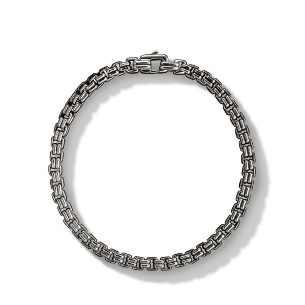 A silver chain bracelet with a box clasp, featuring a pattern of interwoven rectangular links, is displayed against a white background. The intricate design gives the bracelet a textured, layered appearance.