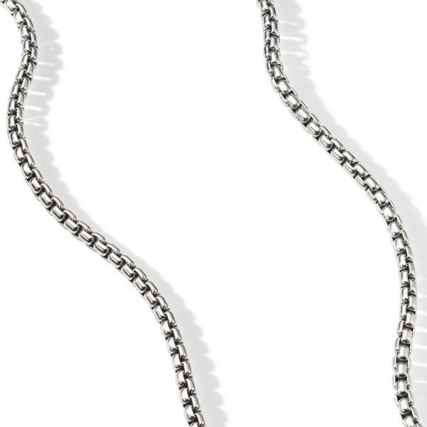 A close-up of a silver chain necklace with interlinked rectangular links, forming a loose, undulating pattern against a white background. The arrangement creates soft shadows that accentuate the contours of the chain.