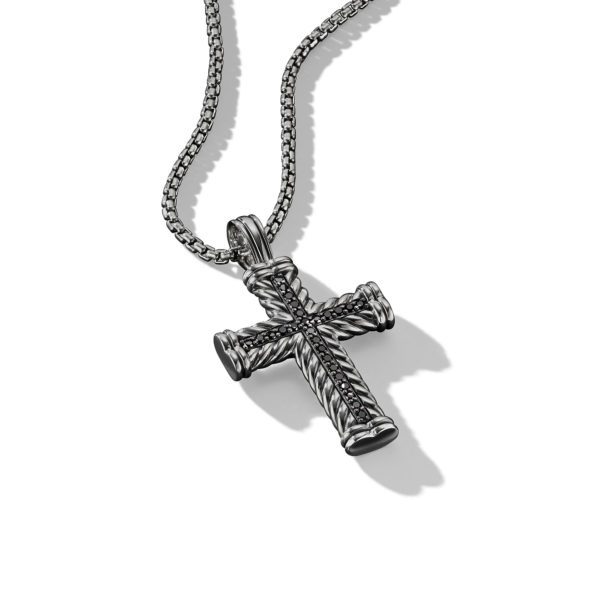 A silver cross pendant with a detailed, twisted rope design hangs from a matching silver chain. The pendant casts a shadow on the plain white background, highlighting the intricate texture of the cross. The chain features a simple, woven pattern.