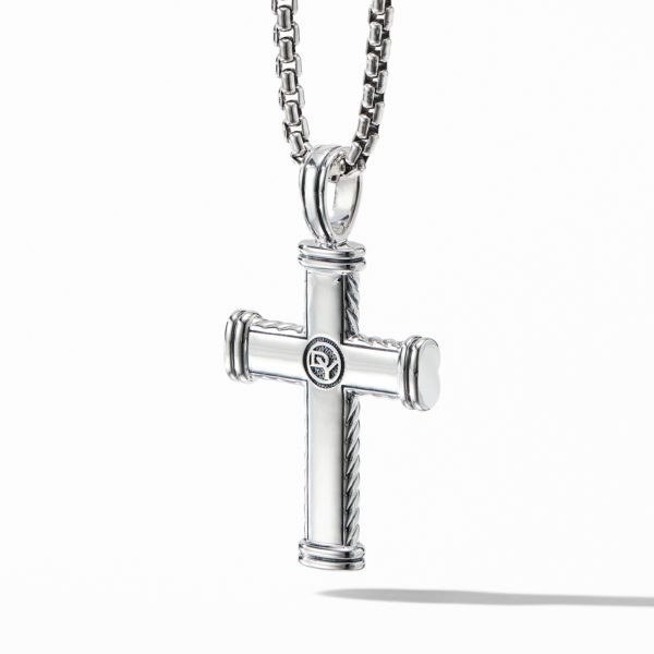 A silver cross pendant hangs from a chain necklace. The cross has a cylindrical design with ridged detailing at the ends of each arm and a small emblem in the center. The background is white, casting a shadow of the cross to its right.