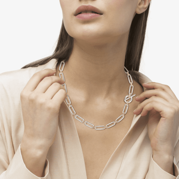 An individual with long hair is wearing a beige blouse and an elegant LAGOS Signature Caviar Two Tone Link Necklace. They are holding the necklace with both hands, gently pulling it forward. Their face is partially visible, showcasing closed lips and a relaxed expression.