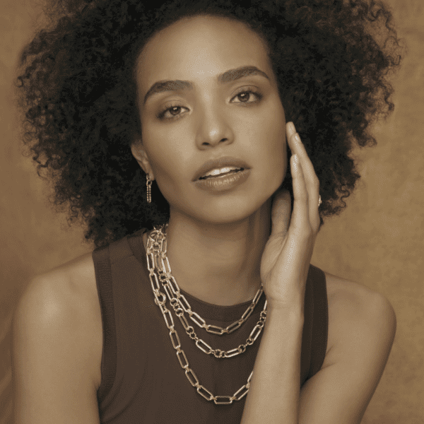 Sepia-toned portrait of a woman with curly hair wearing a sleeveless top and several layered LAGOS Signature Caviar Two Tone Link Necklaces. She lightly touches her face with her hand and looks directly at the camera with a calm expression.