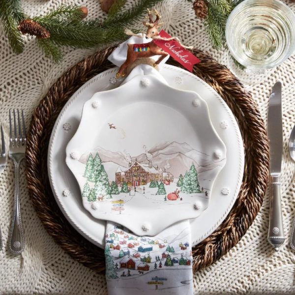 A festive holiday table setting featuring a round woven placemat, a white plate adorned with a snow-covered village scene, and the Juliska Berry & Thread Dessert/Salad Plate - North Pole with its scalloped edge placed on top. A napkin decorated with a winter village is folded beneath the plates. Silverware and a glass are set nearby.