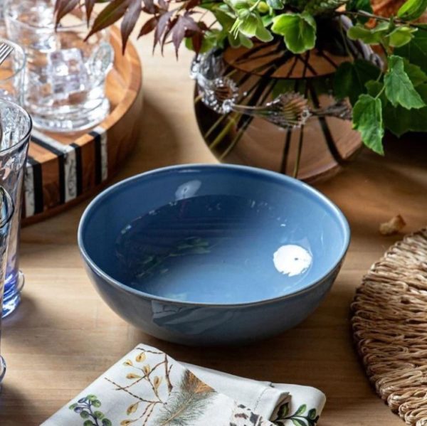 A Juliska Puro Coupe Bowl in Chambray sits on a wooden table next to a folded white cloth with a floral pattern. Surrounding the bowl are a glass, a decorative vase with plants, and a round woven placemat. The table is decorated with various items, creating a cozy atmosphere.