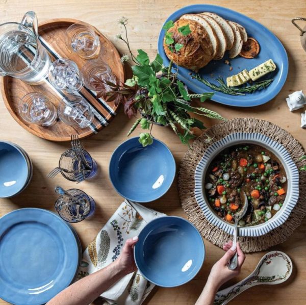 A table set featuring blue ceramic dishes, a wooden tray with glasses and stirring sticks, a bowl of stew with peas and carrots, a plate with sliced bread and cheese, and a vase filled with leafy stems. Hands are placing the Juliska Puro Coupe Bowl - Chambray on the table alongside utensils.