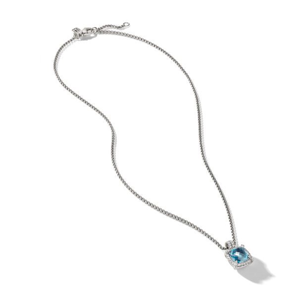 A delicate silver necklace with a fine chain featuring a stunning blue gemstone pendant, surrounded by smaller clear stones, at the end. The necklace is displayed on a white background, showcasing its elegant and simple design.