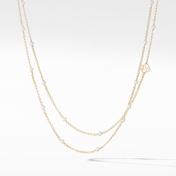 A delicate gold necklace featuring a double-layered chain adorned with small white pearls and a gold monogram pendant near the clasp. The background is smooth and white, highlighting the elegance of the jewelry piece.