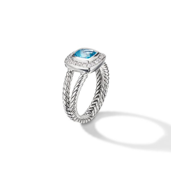 A silver ring featuring a twisted band and a large square-cut blue gemstone at the center, surrounded by small white diamonds. The ring casts a shadow on a plain white background.